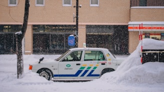 A taxi is parked along a snowy street with a thick layer of snow surrounding it. The background features a brick building with large windows, and snow is gently falling, creating a serene winter scene.