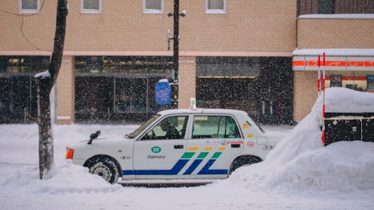 A taxi is parked along a snowy street with a thick layer of snow surrounding it. The background features a brick building with large windows, and snow is gently falling, creating a serene winter scene.