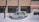 A taxi is parked along a snowy street with a thick layer of snow surrounding it. The background features a brick building with large windows, and snow is gently falling, creating a serene winter scene.