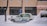 A taxi is parked along a snowy street with a thick layer of snow surrounding it. The background features a brick building with large windows, and snow is gently falling, creating a serene winter scene.