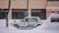 A taxi is parked along a snowy street with a thick layer of snow surrounding it. The background features a brick building with large windows, and snow is gently falling, creating a serene winter scene.
