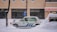A taxi is parked along a snowy street with a thick layer of snow surrounding it. The background features a brick building with large windows, and snow is gently falling, creating a serene winter scene.