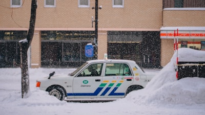 A taxi is parked along a snowy street with a thick layer of snow surrounding it. The background features a brick building with large windows, and snow is gently falling, creating a serene winter scene.