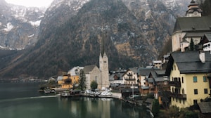 A picturesque lakeside village nestled between a serene body of water and towering mountains. The village features charming, traditional Alpine-style buildings with steep roofs. At the center stands a prominent church with a tall, pointed steeple. The mountains in the background are partially covered in snow, adding to the idyllic, peaceful atmosphere.