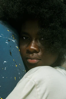 A person with an afro hairstyle is leaning against a surface with paint splatters. The person is wearing a white garment and looking directly at the viewer with a calm expression.