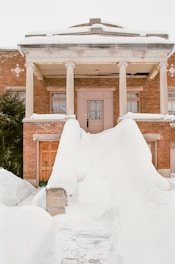 A historic brick building with columns is partially covered in snow. The entrance features large snow drifts leading up to the doorway, and the structure has 'Masonic Temple' inscribed on the facade. The area is serene and blanketed in heavy snowfall.