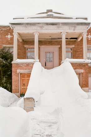 A historic brick building with columns is partially covered in snow. The entrance features large snow drifts leading up to the doorway, and the structure has 'Masonic Temple' inscribed on the facade. The area is serene and blanketed in heavy snowfall.
