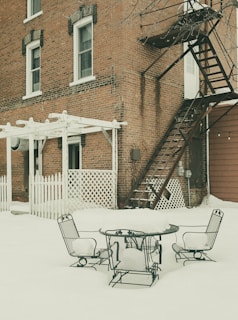 A snow-covered backyard featuring a brick building with two windows and a black metal fire escape staircase. The backyard contains a round metal table and chairs, all dusted with snow, and a white picket fence along with a lattice structure.