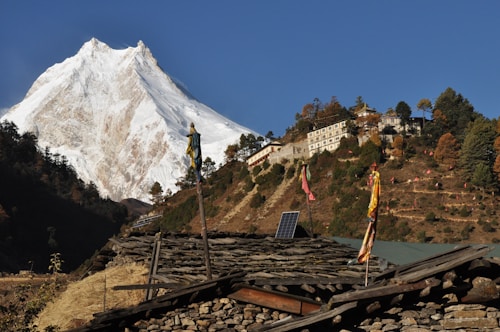 A majestic snow-covered mountain rises under a clear blue sky. Below the mountain, a hillside is adorned with a collection of buildings, probably a monastery, surrounded by evergreen trees. Colorful flags are seen fluttering on poles, and solar panels are visible on the roof of a rustic structure made of stones in the foreground.