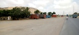 A scene depicting a roadside area with intricately decorated trucks parked in an open space. The trucks have vivid and colorful patterns. There are some trees providing shade and a small structure nearby. A road runs alongside, with a few vehicles, including a motorcycle with a rider.