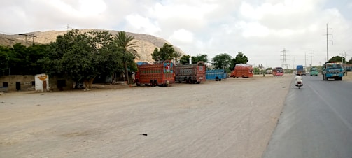 A scene depicting a roadside area with intricately decorated trucks parked in an open space. The trucks have vivid and colorful patterns. There are some trees providing shade and a small structure nearby. A road runs alongside, with a few vehicles, including a motorcycle with a rider.