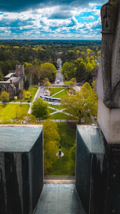 A scenic view from an elevated perspective overlooking a lush green campus with a central statue. The scene includes well-maintained lawns, paved walkways, and a few large historic buildings on the left. Tall trees surround the campus, and a cloudy blue sky stretches across the horizon.
