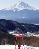A person dressed in red stands facing a traditional red torii gate with the backdrop of Mount Fuji. The landscape includes snow-covered forests and a clear view of the majestic, snow-capped mountain under a pale blue sky.