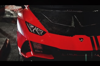 A close-up view of the front of a sleek, red sports car with prominent headlights and a logo on the hood. The lighting creates a dramatic and stylish atmosphere, highlighting the car's aerodynamic design and glossy finish.