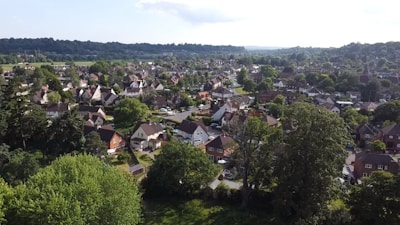 Aerial view of a residential neighborhood with numerous houses surrounded by lush greenery. The houses are predominantly detached with pitched roofs, and there's a dense coverage of trees and lawns. The landscape extends to a horizon of hills, giving a sense of a suburban area nestled within nature.