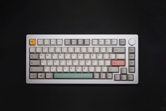 A mechanical keyboard with a compact layout sits against a black background. The keycaps are mostly gray with some accentuated keys in different colors, including a pink Enter key, a light green spacebar, and an orange Escape key. The keyboard has a metallic finish and features both alphanumeric keys and function keys. A large rotary knob is located in the upper right corner.