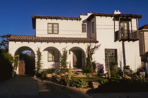 A two-story house with a Mediterranean architectural style featuring white stucco walls and a red-tiled roof. The front of the house has arched doorways and a small balcony on the second floor. The garden is landscaped with various shrubs and plants, and a stone pathway leads to the entrance. The scene is lit by late afternoon sunlight, casting long shadows.