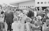 A woman in formal attire, holding a bouquet of flowers, is interacting with a group of children in what appears to be a public event. She is surrounded by several adults, some in formal suits, while a crowd gathers around the scene. A modernist building with circular windows is visible in the background, along with some trees on the left side of the image.