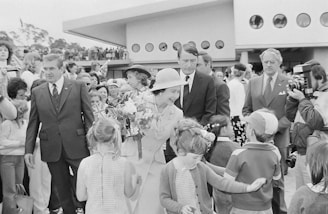 A woman in formal attire, holding a bouquet of flowers, is interacting with a group of children in what appears to be a public event. She is surrounded by several adults, some in formal suits, while a crowd gathers around the scene. A modernist building with circular windows is visible in the background, along with some trees on the left side of the image.