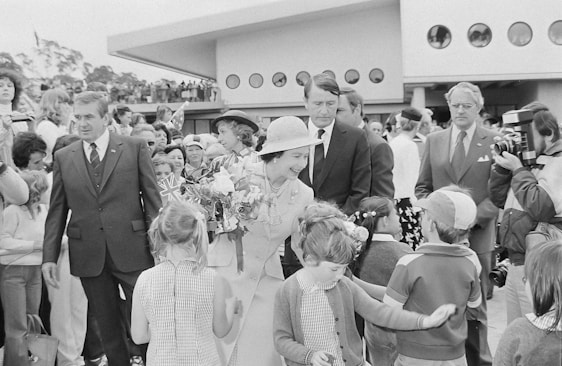 A woman in formal attire, holding a bouquet of flowers, is interacting with a group of children in what appears to be a public event. She is surrounded by several adults, some in formal suits, while a crowd gathers around the scene. A modernist building with circular windows is visible in the background, along with some trees on the left side of the image.