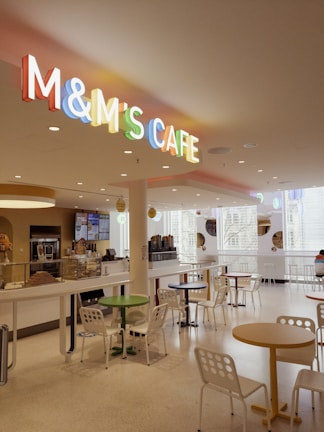 A modern, well-lit cafe interior with a colorful M&M's Cafe sign overhead. The space features a variety of round tables and chairs with a white and minimalistic design. Multiple windows allow natural light to fill the cafe, and a counter area is visible with a menu board and beverage machine.