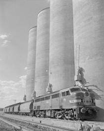 A black-and-white image featuring a train positioned alongside a series of large grain silos. The scene conveys an industrial setting with the train and the massive cylindrical structures dominating the landscape.