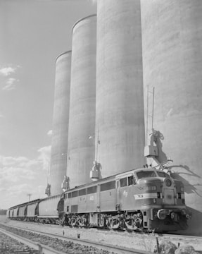 A black-and-white image featuring a train positioned alongside a series of large grain silos. The scene conveys an industrial setting with the train and the massive cylindrical structures dominating the landscape.