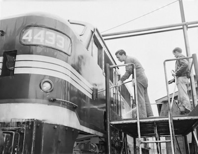 Two individuals are seen cleaning the exterior of a locomotive with the number 4433 displayed on the front. They are standing on a platform using hoses to wash the surface. The locomotive sports a vintage design with stripes on its metal body, and the atmosphere appears industrious and focused.