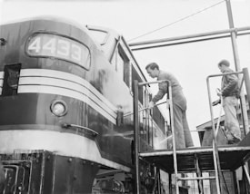 Two individuals are seen cleaning the exterior of a locomotive with the number 4433 displayed on the front. They are standing on a platform using hoses to wash the surface. The locomotive sports a vintage design with stripes on its metal body, and the atmosphere appears industrious and focused.