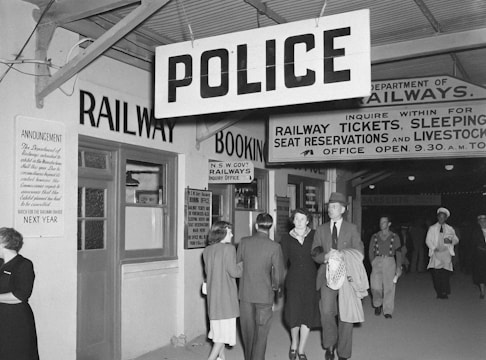 A vintage black and white photo of a railway station interior, featuring people walking beneath large signs for 'Police' and various railway services. The station appears busy, with business attired individuals and a policeman visible.