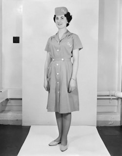 A woman stands in a professional pose, wearing a uniform dress with buttons down the front and a matching hat. The outfit has the letters 'J L' embroidered on it. The setting is a simple indoor backdrop with minimal decor.