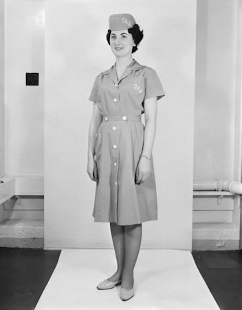 A woman stands in a professional pose, wearing a uniform dress with buttons down the front and a matching hat. The outfit has the letters 'J L' embroidered on it. The setting is a simple indoor backdrop with minimal decor.