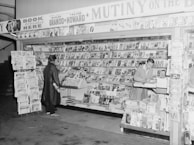 A black and white image of a vintage bookstore or magazine stand. The shelves are filled with various magazines and books. Two individuals are present; one person is a customer browsing the selection, while another is behind the counter, possibly organizing the books. Signage above highlights a book titled 'Mutiny on the Bounty' featuring Marlon Brando and Trevor Howard, suggesting a specific time period. The setting appears organized and inviting, typical of mid-20th-century print media displays.