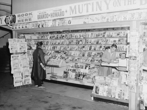 A black and white image of a vintage bookstore or magazine stand. The shelves are filled with various magazines and books. Two individuals are present; one person is a customer browsing the selection, while another is behind the counter, possibly organizing the books. Signage above highlights a book titled 'Mutiny on the Bounty' featuring Marlon Brando and Trevor Howard, suggesting a specific time period. The setting appears organized and inviting, typical of mid-20th-century print media displays.