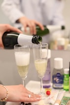 Two champagne glasses are being filled with sparkling wine from a green bottle with a white label. One person holds the bottle, and another hand with a bracelet and a ring is resting on the table. Various beauty products and a small pom-pom are visible on the table, adding a casual yet elegant context.
