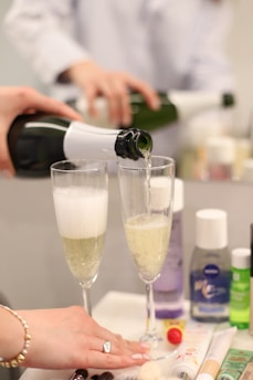 Two champagne glasses are being filled with sparkling wine from a green bottle with a white label. One person holds the bottle, and another hand with a bracelet and a ring is resting on the table. Various beauty products and a small pom-pom are visible on the table, adding a casual yet elegant context.