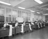 A spacious office room with high ceilings and fluorescent lighting. Several men are seated at wooden desks with typewriters, each producing long sheets of paper that spill over the side. The room has large windows and various office supplies scattered across the desks.