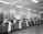 A spacious office room with high ceilings and fluorescent lighting. Several men are seated at wooden desks with typewriters, each producing long sheets of paper that spill over the side. The room has large windows and various office supplies scattered across the desks.
