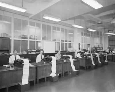 A spacious office room with high ceilings and fluorescent lighting. Several men are seated at wooden desks with typewriters, each producing long sheets of paper that spill over the side. The room has large windows and various office supplies scattered across the desks.