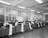 A spacious office room with high ceilings and fluorescent lighting. Several men are seated at wooden desks with typewriters, each producing long sheets of paper that spill over the side. The room has large windows and various office supplies scattered across the desks.