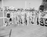 A group of women in swimsuits are standing together on a platform or stage in an outdoor setting. They appear to be participating in a beauty pageant or similar event. In the background, there are several buildings and trees, with some people observing the scene. The image is in black and white, suggesting it may be from a historical period.