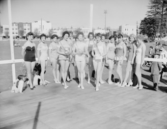 A group of women in swimsuits are standing together on a platform or stage in an outdoor setting. They appear to be participating in a beauty pageant or similar event. In the background, there are several buildings and trees, with some people observing the scene. The image is in black and white, suggesting it may be from a historical period.