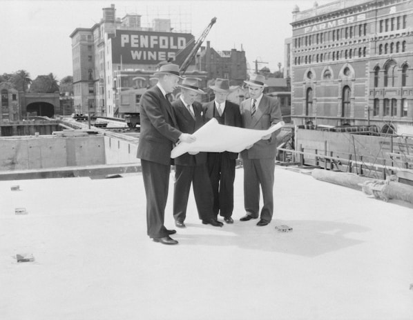 Five men in suits and hats are standing together on a construction site, examining a large blueprint. Behind them, there are two prominent buildings with signs that read 'Penfold Wines' and 'Farmers'. Construction equipment and materials are visible in the background.