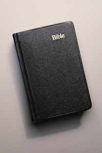 A black leather-bound Bible with 'Bible' written in gold lettering on the cover, lying on a flat, neutral-colored surface.
