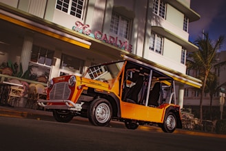 A bright orange, vintage-style electric car is parked in front of a classic art deco building, with 'The Carlyle' signage visible above. The setting includes tropical elements, such as palm trees and outdoor café seating. The overall ambiance hints at a sunny, coastal location, possibly during the late afternoon or early evening.