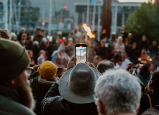 A crowded outdoor event with many people gathered, some wearing colorful clothing and hats. In the foreground, a person is taking a photo or video with a smartphone, capturing a flame. The background includes blurred figures, suggesting a lively and dynamic atmosphere.