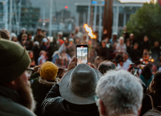 A crowded outdoor event with many people gathered, some wearing colorful clothing and hats. In the foreground, a person is taking a photo or video with a smartphone, capturing a flame. The background includes blurred figures, suggesting a lively and dynamic atmosphere.