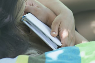 A close-up of a hand holding a book with a violet cover, surrounded by soft pink and gray lighting.