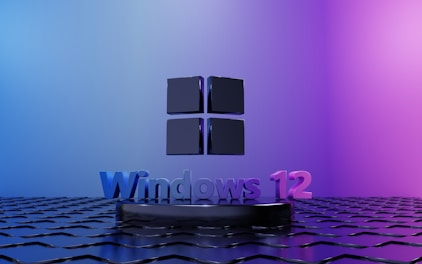 A stylized black Windows logo is positioned above a textured black surface with a metallic sheen. Below it, the text 'Windows 12' is displayed in a gradient of blues and purples. The background transitions from light blue on the left to pinkish-purple on the right, creating a dramatic and modern look.