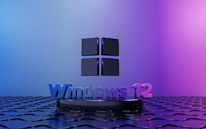 A stylized black Windows logo is positioned above a textured black surface with a metallic sheen. Below it, the text 'Windows 12' is displayed in a gradient of blues and purples. The background transitions from light blue on the left to pinkish-purple on the right, creating a dramatic and modern look.